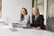 © fizkes - Cheerful younger and older female business colleagues sitting at workplace table in office, looking away, smiling, laughing, working at laptop together, promoting professional communication