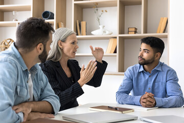 Diverse business team of serious colleagues discussing project at meeting table, talking, sharing ideas for brainstorming. Senior manager woman, consultant, mentor speaking to coworkers