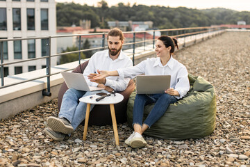 Wall Mural - Two coworkers sitting on rooftop workspace collaborating using laptops. Casual setting, outdoor work environment, enhancing teamwork and productivity. Relaxed atmosphere promotes open communication.