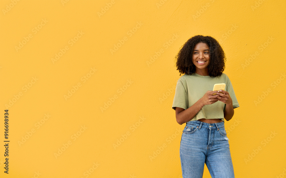 Young happy African gen z teen girl standing at yellow wall looking at ...