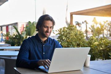 Happy young business man entrepreneur using laptop sitting outdoors with coffee. Smiling guy student or professional worker looking at computer in cafe elearning, hybrid working, searching job online.