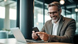 © halo - An office worker is smiling while using his phone, sitting at an open desk with a laptop in front of him.