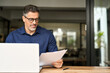 © insta_photos - Busy middle aged professional business man using laptop computer reading document at work. Senior mature male manager executive working on laptop checking paper financial report in office. Copy space.