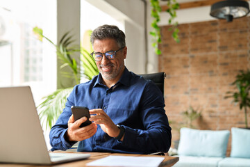 Happy middle aged businessman using mobile phone working in office. Busy smiling 50 years old man executive holding smartphone technology looking at cellphone making bank transaction payment in app.