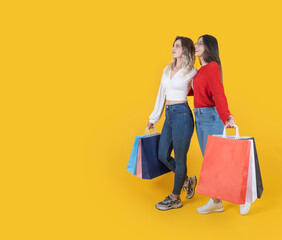 Full body length side view two beautiful fit caucasian 20s young women wear casual slim jeans and shirt carry many shopping bags walking looking aside copy space over isolated yellow studio background