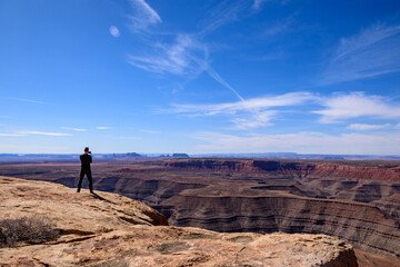 A man stands on an overlook at Muley Point in Utah, taking photos of the expansive and breathtaking desert landscape under a blue sky. This image captures adventure, travel, and natural beauty - USA