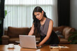 © wattana - Young woman in professional attire stands at a desk, intently focused on a laptop screen in a cozy, modern office environment.
