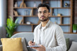 © Tetiana - Portrait of a young Indian man sitting on the sofa at home, holding a notebook and a pen in his hands, looking seriously and confidently at the camera