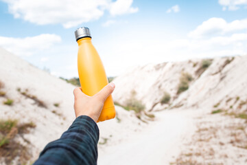 Wall Mural - Male hand holding steel thermo water bottle of yellow color on background of sand hills.