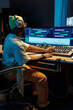 © pressmaster - Music producer working in a modern studio with advanced sound equipment, wearing headphones and operating a large mixing board, surrounded by screens displaying audio tracks