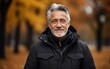 © imagineRbc - A man with a beard and gray hair is smiling in a black jacket. He is wearing a scarf and he is enjoying the autumn weather