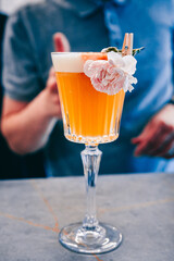 Wall Mural - A close-up of a cocktail in a stemmed glass, garnished with a pink flower, placed on a surface. An out-of-focus person is visible in the background, adding depth to the scene