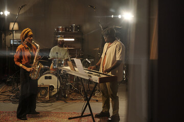 Musicians jamming in studio with saxophonist, drummer, and keyboardist engaged in passionate performance under warm lighting creating an intimate atmosphere