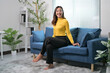 © Parichat - Woman in Yellow Sweater Sitting on Blue Sofa in Modern Living Room with Indoor Plants and Natural Light