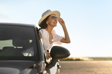 Wall Mural - Smiling young woman in sunglasses with hat leaning out of car window outdoors. Enjoying trip