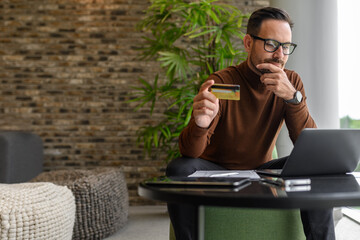 Smiling male accountant paying bills with credit card over computer while sitting at table in office