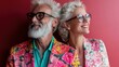© Vuvimages - A vibrant image showcasing an elderly couple dressed in eye-catching, colorful floral attire, standing against a striking red background, exuding charisma and individuality.