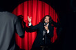 © Parilov - Smiling man speaking to microphone performing in comedy show open mike stand up on stage with red curtains