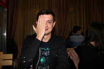 Man Playfully Covering One Eye in a Social Setting
Man in casual attire covering one eye with his hand, sitting in a cozy social setting.