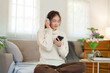 © Pichsakul - Young asian women in sweater wear headphone and listen music with enjoying on smartphone while sitting on the couch to relaxation and doing activity for spending time with slow life lifestyle at home