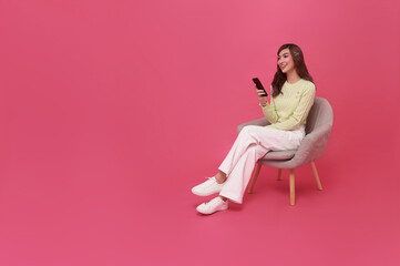 Wall Mural - Full length beautiful Asian teen woman using smartphone and sitting on chair isolated on pink copy space background. People lifestyle Technology Concept.