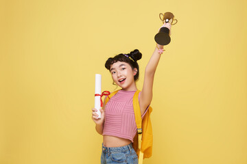 Canvas Print - Happy graduate asian schoolgirl with a diploma and trophy shows a gesture of victory and success isolated on a yellow background.