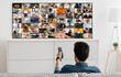 © Prostock-studio - A man is sitting on a couch and watching television, holding the remote control in his hand. The TV screen is showing a grid of images, each depicting a different scene.