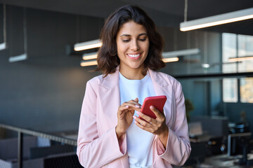 European business woman manager using cell phone mobile app standing indoors in office. Latin Hispanic young female businesswoman entrepreneur focused working on smartphone, do a call, texting message