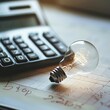 © markusmiller - Small bulb next to a calculator on a white board, illustrating the intersection of illumination