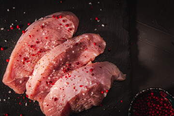 Wall Mural - Raw turkey steaks with spices on a black mica board on a gray background with a bowl of pink pepper. Copy a space