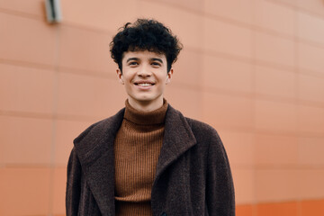 Wall Mural - Fashion portrait of a young man with curly hair standing in front of an orange wall, smiling and wearing a brown coat and turtleneck