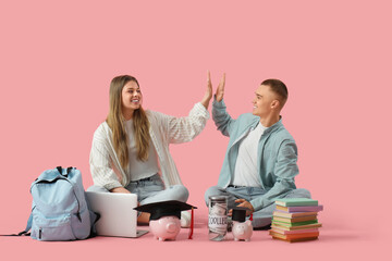 Canvas Print - Young students giving high-five gesture and sitting with piggy bank, books, backpack and laptop on pink background. Concept of savings for education