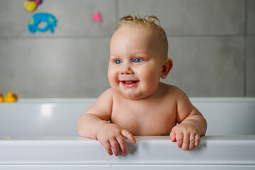 Wall Mural - A smiling baby is standing in a white bath with water. Baby bathing in water. Personal hygiene of the baby.