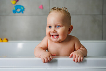 Wall Mural - A smiling baby is standing in a white bath with water. Baby bathing in water. Personal hygiene of the baby.