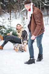 Beautiful young loving couple having fun spending winter vacation in a countryside cottage, having fun riding on sled on the snow. Wearing warm clothes, scarf, knitted sweater. Holiday cozy atmosphere