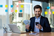© Liubomir - Businessman in suit smiling while holding cash and using smartphone at office desk. Man working on laptop, making online transactions, managing finances. Concept of success, financial growth, business