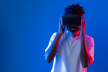 Surprised young African American looking through VR holding interesting object hologram on pink blue neon wall at metaverse world connecting digital futuristic technology virtual reality. Contrivance.