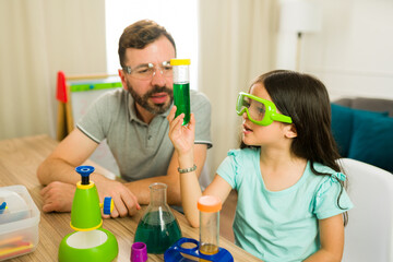 Father and his daughter wearing safety glasses while homeschooling at the dining table, conducting a science experiment with a test tube