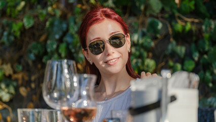 A beautiful young red-haired woman smiling while posing with a drink in her hand at a picnic table on a summer day outdoors