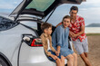 © InfiniteFlow - Family vacation trip traveling by the beach with electric car, lovely family sit on the trunk, charging EV car battery with green and sustainable energy. Family travel and eco-friendly car. Perpetual