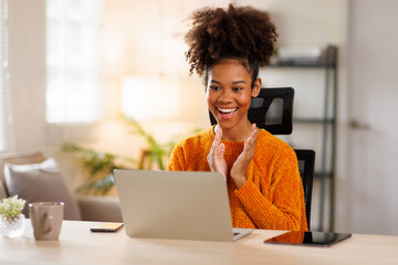 Female digital nomad smiling happily as she types on her laptop on sofa. Afro haired woman dressed in casual clothing and working online in a relaxed environment.