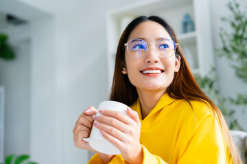 Young asian woman wearing eyeglass sitting on sofa and holding mug in living room at home, drinking milk or tea in the morning. Healthy and refresh mood