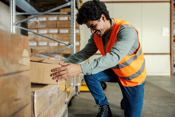 Wall Mural - Smiling diverse warehouse worker kneeling at facility and taking box from shelf and preparing shipment.