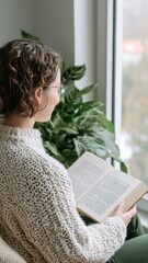 young woman reading book by window in cozy home interior with plant