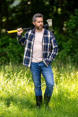 Wall Mural - Lumberjack with serious face carries axe outdoor. Lumberjack worker in the forest with axe. Strong man lumberjack with an ax in a plaid shirt. Lumberjack or logger in forest.