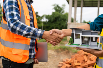 Two male architects, wearing hard hats and safety vests, discuss construction design while holding hands.   contractor, focusing on the house structure, iron framework, brickwork, and roofing.