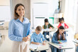 © Home-stock - Happy friendly female teacher smiling at camera during lesson at elementary school while pupils writing in copybooks on background in classroom