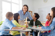© Home-stock - Happy diverse kids listening their teacher while sitting at desks in classroom, looking at woman and smiling