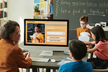 Back view of boy using computer in school with female teacher explaining smart learning course to student