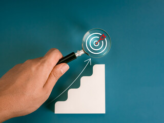 Wall Mural - Business market trends, investment goal and success, marketing search tool concept. Target icon focus in magnifying glass in man's hand and white stair graph steps with rise arrow on blue background.